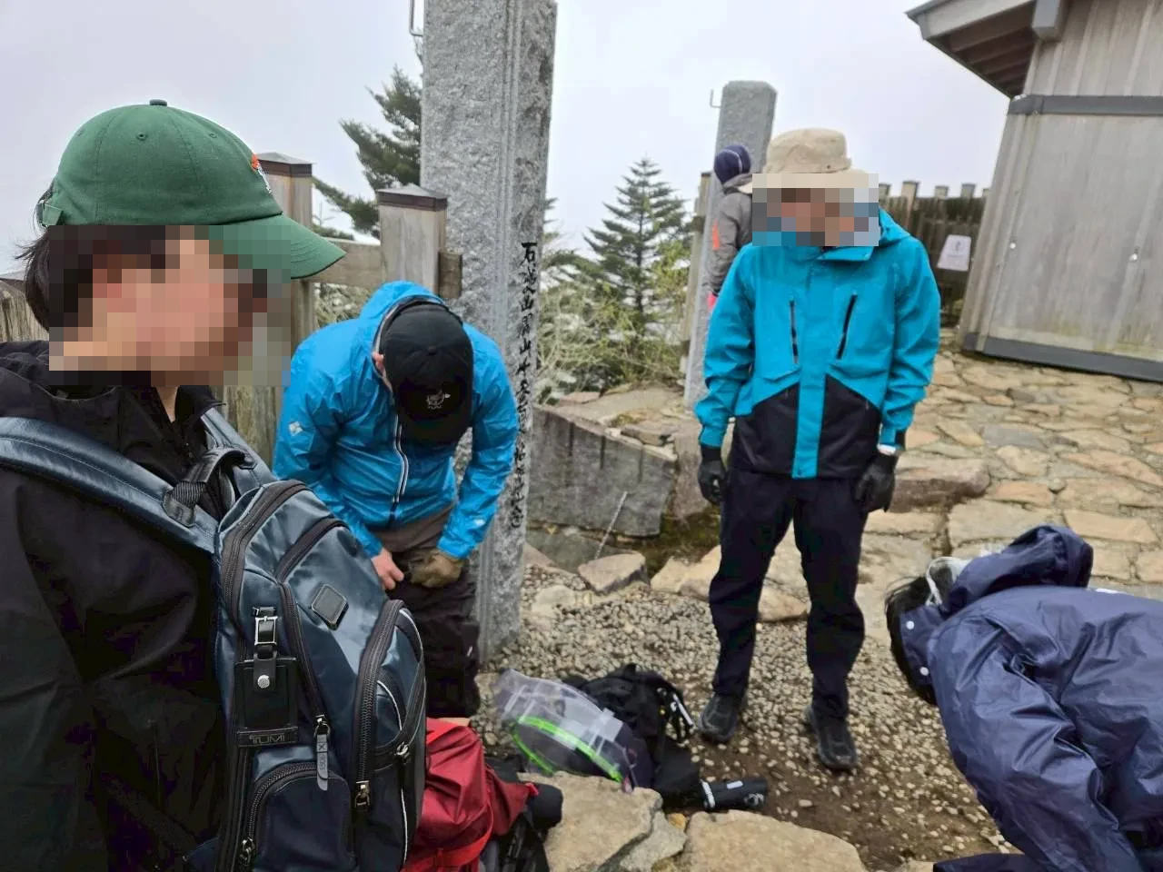 山頂付近で装備を確認している登山者たち。霧の中、電波塔の柱のそばで立ち止まっている。